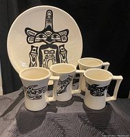 Front view of 4 cups with black Indigenous-style artwork and 1 plate with central black figure on white glaze. Cups have squared handles.