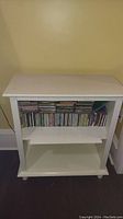 Front view of white wooden bookcase with two shelves, containing several CDs on the top shelf, lower shelf empty
