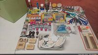 Wide layout photo showing assorted arts and crafts supplies including a variety of paint tubes, markers, inks, rubber stamps, broken china pieces, scissors, and pliers arranged on a white surface and bin.
