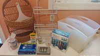 Wide shot of assorted bathroom items on a white surface near window: three white plastic file holders, metal shower caddy, brown wicker basket, hair clipper box, razor blade pack, glass jars, toothpaste, small container.