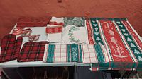 Full assortment of twelve kitchen and decorative linens, oven mitt, pot holder, and Christmas ribbons laid out on a white table against a red wall.