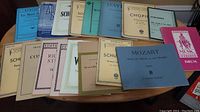 Top-down photo of 18 classical music songbooks spread on wooden table, showing titles for Mozart, Schubert, Chopin, Haydn among others.