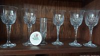 Five crystal wine glasses with diamond cut pattern, one smaller plain glass, and small herb container placed on wooden shelf