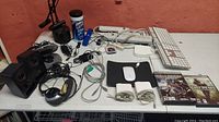 View of entire lot including desk lamp, keyboards, mouse, charger cords, speakers, headphones, portable CD player, game discs on a white table against a brown wall