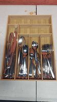 Flatware including knives, multiple sizes of spoons, and forks organized in a wooden compartmentalized tray on a white surface.