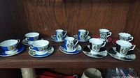 Photo showing ten porcelain tea cups and matching saucers arranged on a wooden shelf, featuring varied classical art designs and gold accents.