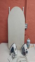 Two irons, small ironing board standing vertically, ironing spray can on table surface against wall