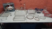 Full view of assorted glass kitchenware items spread on table, including Pyrex glass containers, Anchor Hocking glassware, silver plated serving tray with warming stand and lid in box, measuring cups, plates, and glass bottle.