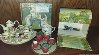 Grouping of two miniature tea sets and two decorative tea time themed boxes shown on wooden surface.