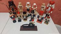 Front view of 15 nutcracker figurines displayed on a white surface against a red wall, showing variety in size and style.