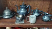 Full view of the pewter tea set including teapot, sugar bowl, creamer, lidded serving dish, tin mug, and decorative cup on tray.