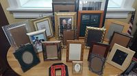 View of all 16 picture frames of various sizes, styles and colors arranged on a wooden table in front of a window. Some frames include pictures, others are empty.