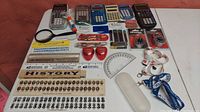 Photo shows a variety of school supplies spread on a white table with orange background. Items include calculators, rulers with historical timeline, pencil sharpeners, protractor, tape measures, magnifying glass and small supplies such as pencil lead and erasers.