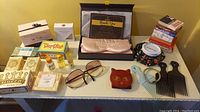 Overview of assorted women's personal items displayed on white table including soaps, sunglasses, small books, hair picks, and an eye mask.
