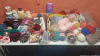 Large table filled with many skeins of yarn in assorted colors and textures along with crocheting needles and other supplies.