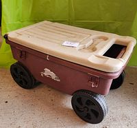 Closed view of brown plastic garden cart wagon with cream lid and black wheels on concrete floor against green background.