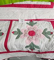 Close-up view of the handmade single quilt showing detailed flower appliqué design with pink petals, green leaves, and red center, bordered by red fabric on a white background.