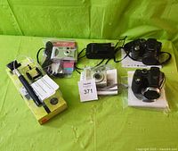 Group photo of all 6 items: Canon Sprint, FujiFilm FinePix S, FujiFilm FinePix S700, Canon PowerShot A510, Ixla Super Pro 640, and Hype selfie stick with packaging on green tablecloth.