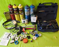 Overview of tools, gas cans, solder rolls and other soldering supplies arranged on a green surface, including a hammer and soldering iron in case.