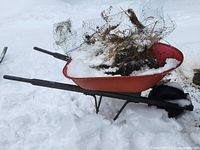 Red metal wheelbarrow with black handles and a solid flat-free tire shown outside with snow and some dried plant debris inside the tray.
