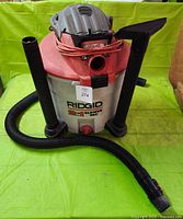 Front view of RIDGID 2-in-1 wet/dry vacuum showing the red top and silver body, with the power cord wrapped around the top handle. Attached to the vacuum is the corrugated long hose and two straight black extension tubes.