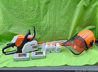 Layout of chainsaw, helmet, small oil bottles, spare chain boxes and chain oil jug on green backdrop.