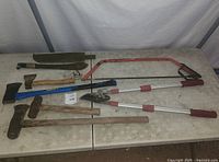 Photo showing a set of eight wood cutting tools on a table including axes, saws, hammer, maul, and loppers with wooden handles and metal blades.