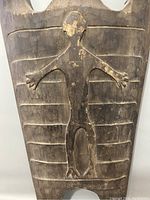 Close-up of the central carved human figure on the wooden shield displaying the convex relief and horizontal lines.