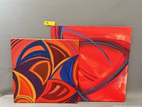 Two modern abstract geometric oil paintings on canvas leaning against a wall. The left painting has vibrant orange, blue, red and purple curved geometric forms. The painting on the right is mostly red with black and blue sweeping arcs.