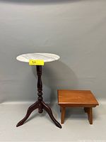 Photo showing a round white marble top stand with a dark turned wooden pedestal and tripod legs next to a small wooden bench with a rectangular top.