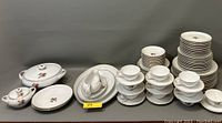 Full display of white porcelain dinnerware with pink rose pattern arranged in groups including plates, cups, bowls, and serving dishes