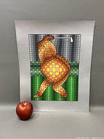 Front view of the unframed serigraph print showing the colorful abstract golfer figure with yellow, red, and green squares and a golf club in a grid background. An apple is placed beside it for scale.