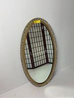 Full view of vintage ornate gilt oval wall mirror leaning against a white wall with a wooden frame featuring detailed carved gold gilt floral and scroll motifs.