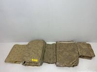 Photo showing seven folded pieces of vintage upholstery fabric with damask and floral patterns and gold-beige colors, arranged side by side on a white surface against a plain wall.