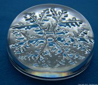 Clear glass paper weight with detailed embossed snowflake design on a dark blue background, showing slight reflection and rainbow effect on edges.