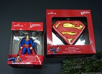 Photo showing both Hallmark Superman Christmas tree ornaments in original packaging, one a figurine and one a glass shield logo.