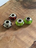 Four assorted glass and sterling silver bead charms, colors black and white, red and black, and green and white dots, displayed on a wooden surface.