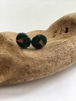 Close-up photo of pair of round stud earrings with green malachite and red coral mosaic in silver base resting on carved wood stand.