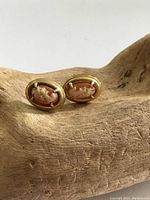 Front view of two oval antique yellow metal stud earrings with hand carved shell white cameo on peach background, placed on light wood surface