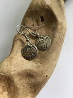 Pair of silver North West Coast Native earrings resting on a carved wooden display, showing one earring close up with visible indigenous art design and hook.