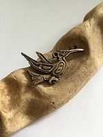 Copper coloured pewter hummingbird brooch resting on a textured beige surface, showing detailed engraved patterns and form of a hummingbird in North West Coast style.
