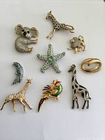 Top view photo showing group of eight animal-themed rhinestone brooches including koala, giraffes, lion, bird, starfish and an abstract brooch.