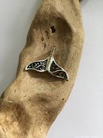 Photo of the sterling silver whale tail pendant placed on a piece of wood, showing detailed swirl designs and colorful inlay on the tail.