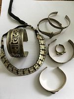 Photo of all jewelry pieces laid out on white background showing the large cuff bracelet with Tibetan script, the stone-inset necklace, multiple other cuff bracelets, and the open-ended ring with colored stones.