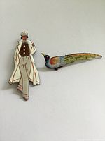Front view of two vintage brooches, one depicting a man in a cape and the other a colorful bird, arranged on a plain surface.