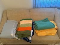 View of multiple folded blankets on a beige couch including a light blue fabric blanket with satin trim, a crocheted blanket with green, cream, and orange stripes, solid green and yellow blankets.