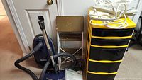 Photo showing blue Kenmore vacuum cleaner, metal folding step ladder, yellow and black plastic drawer storage unit with items on top in a room corner