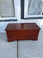 Front view of the reddish-brown rectangular cedar chest on concrete with white-framed windows in background
