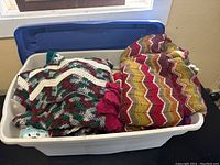View of a white plastic storage bin showing two crochet blankets, one burgundy, gold, and white in chevron pattern, and another with block colors.