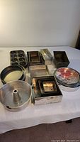 Overview of bakeware lot on table showing variety of metal pans including round, square, rectangular, bundt, and muffin tins with visible wear
